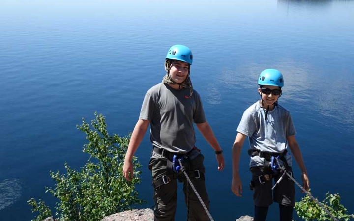 Two people wearing safety gear are secured by ropes as they lean back over a blue body of water. 